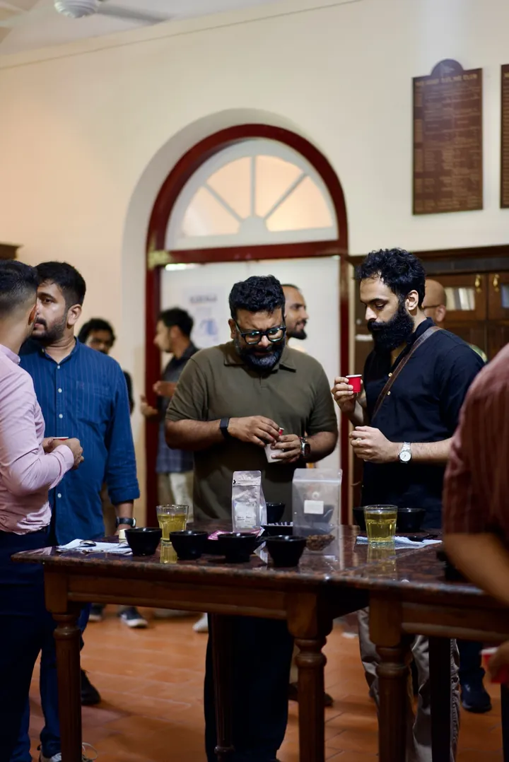Guests tasting coffee from small red cups beside the Kochi cupping table.